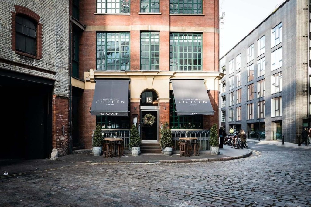 Exterior view of Jamie Oliver's Fifteen Restaurant and Bar, with black awnings, situated in a restored brick building on a cobblestone street, illustrating urban redevelopment and the blending of historical architecture with modern gastronomy.