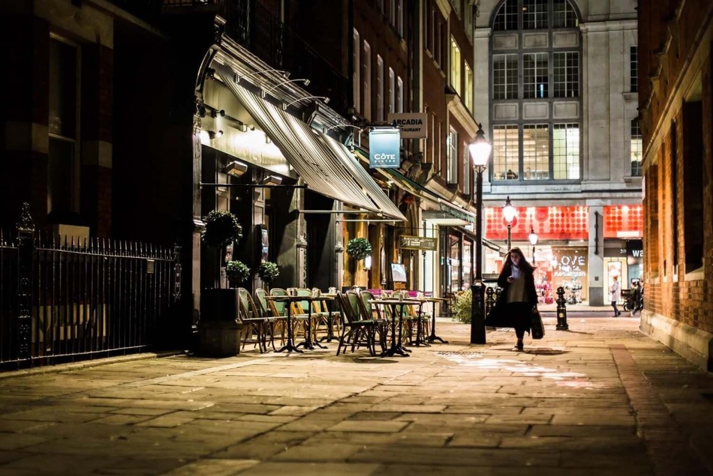 Nighttime scene of an atmospheric London alley with the warmly lit facade of Côte Brasserie, empty outdoor seating ready for diners, and a solitary figure walking by, evoking the city's intimate dining culture.