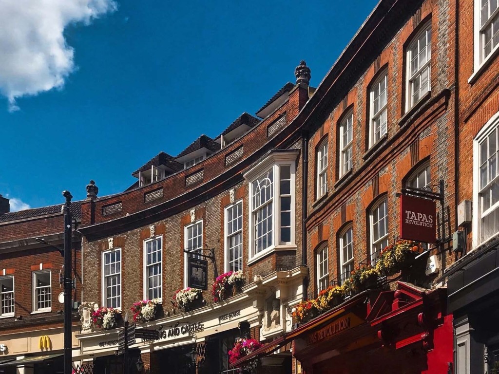 Charming street view of traditional British architecture with red brick buildings adorned with flower baskets, featuring the sign for Tapas Revolution, under a clear blue sky, capturing the essence of urban cultural and gastronomic offerings.