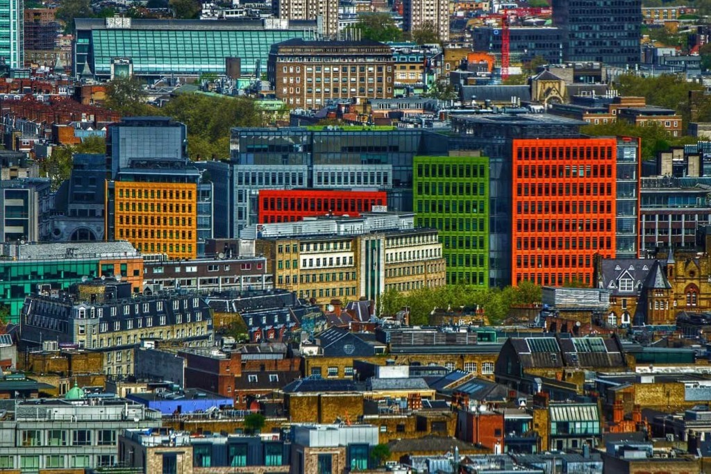 High-angle view of London's diverse architectural landscape, showcasing a vibrant patchwork of historic and modern buildings with colorful facades in orange, red, and green, illustrating the city's dynamic urban fabric and development.