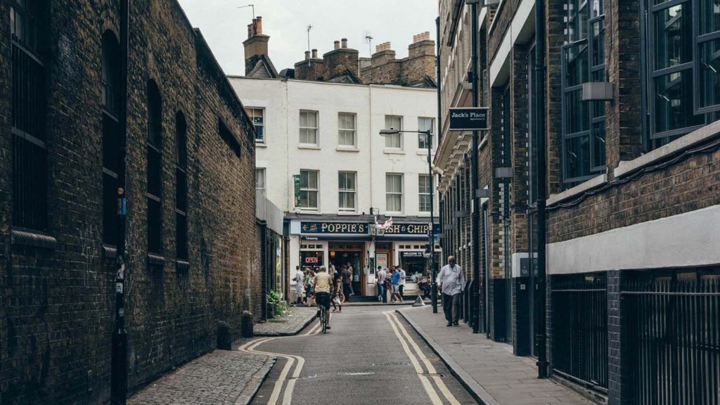 Quaint alleyway in London leading to Poppie's Fish & Chips, a traditional British eatery, with pedestrians and a cyclist adding life to the scene, flanked by historic brick buildings, embodying classic urban street life in the UK.