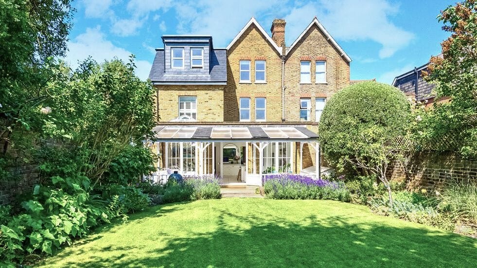 Classic Victorian brick house with a charming conservatory, overlooking a lush garden with well-trimmed hedges and flowering plants, under a clear blue sky, reflecting a seamless blend of historical elegance and modern comfort.