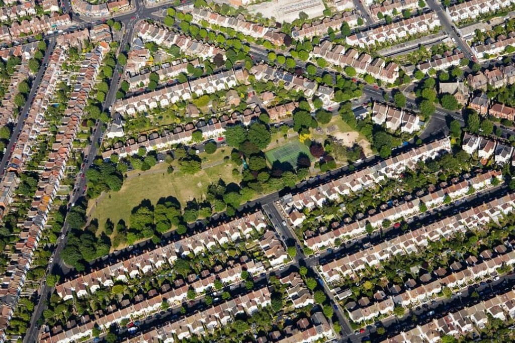 Aerial view of a suburban neighbourhood with rows of identical houses and manicured lawns, divided by a network of roads and a central green park area, showcasing residential planning and community layout from a bird's-eye perspective.
