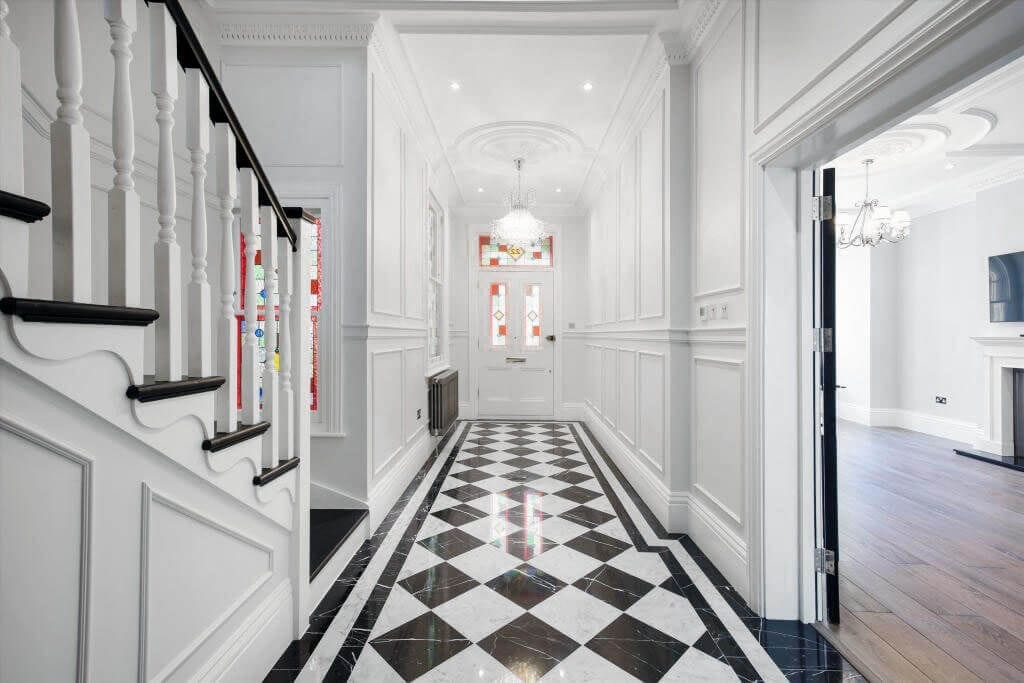 Elegant entryway with a classic black and white checkered floor, leading to a spacious living room with dark hardwood floors, featuring detailed wainscoting, a white staircase with black accents, and a bright, welcoming atmosphere enhanced by natural light.