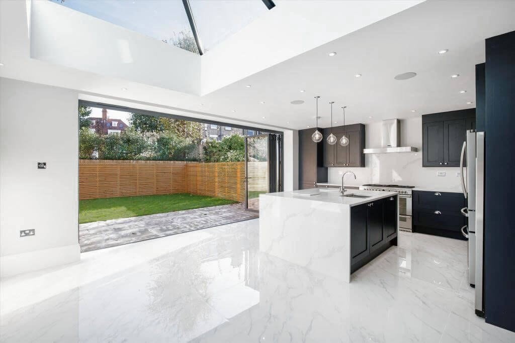 Luxurious modern kitchen with high-gloss marble flooring, featuring a large central island, sleek black cabinetry, and stainless-steel appliances, well-lit by natural light from overhead skylights and floor-to-ceiling sliding glass doors opening to a manicured garden.