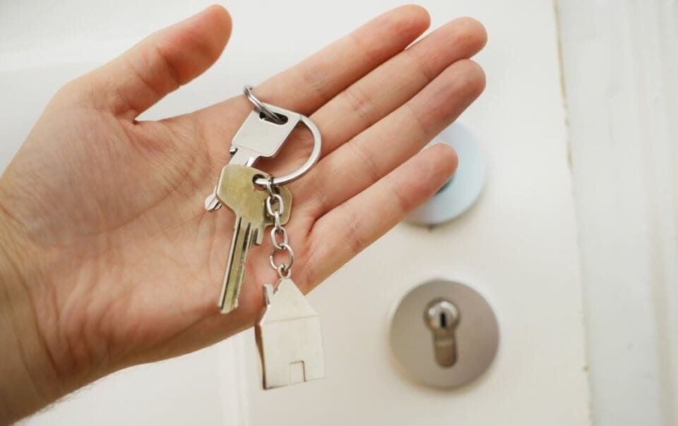 New homeowner holding in front of their front door the keys to their forever family home on their left index finger with the little house keychain resting on their palm