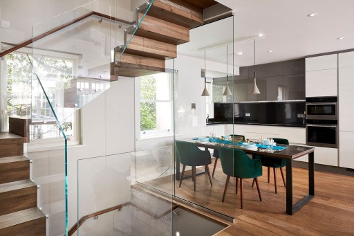 Shiny glass stairwell landing on the same floor at a medium sized wooden formal dinner table and the monochromatic kitchen in the background
