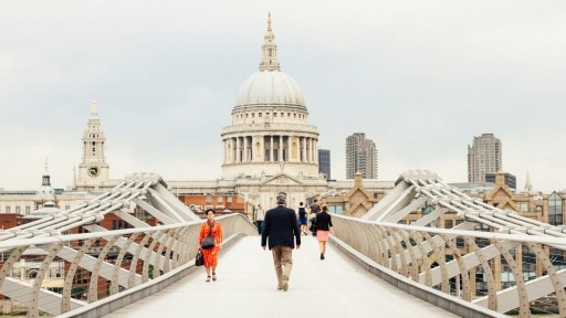 View of St. Paul's Cathedral from Millennium Bridge in Central London, with people walking on the bridge, showcasing the mix of historic architecture and modern infrastructure.
