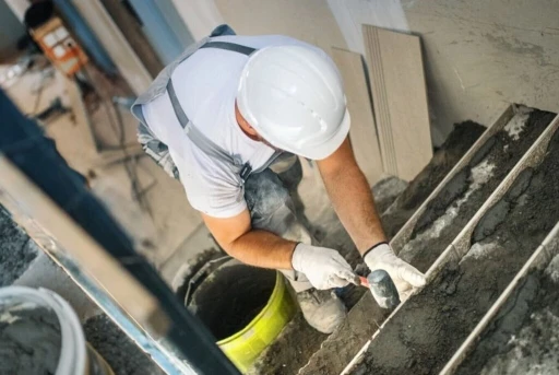 Builder wearing a hard hat and gloves laying floor tiles during a house renovation project.