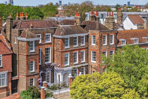 Close-up view of Chiswick's charming red brick townhouses with white sash windows, classic British architectural details, wisteria blooms, and terracotta chimney pots, set against a backdrop of green treetops and urban skyline.