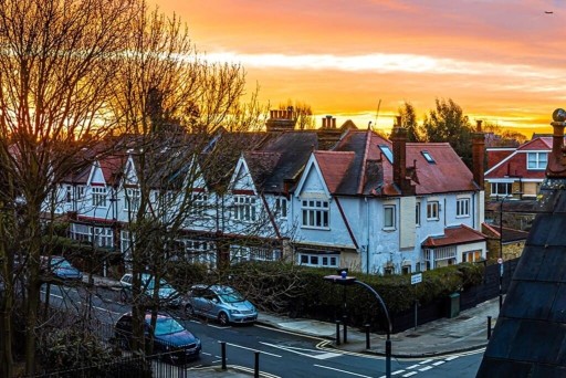 Dawn breaks over a peaceful Chiswick residential street, casting a warm glow on quaint white houses with gabled roofs and cars parked along the road, showcasing a quintessentially British suburban scene.