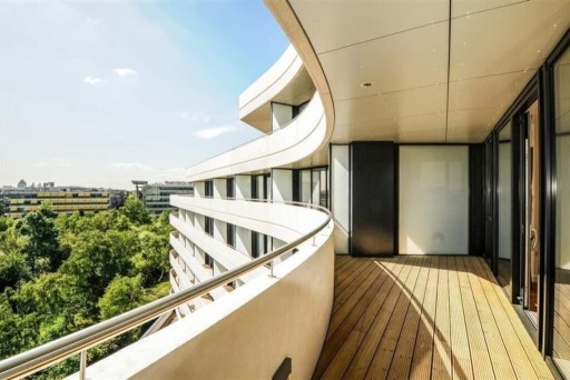 Modern apartment balcony with curved design, wooden flooring, and sleek glass balustrades overlooking a lush green park, reflecting upscale urban living and contemporary architecture.
