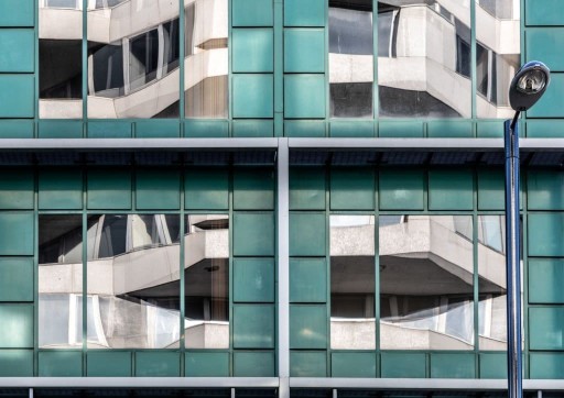 Four large rectangular windows with reflective film mirroring the opposite large new build development encased with modern teal tile cladding
