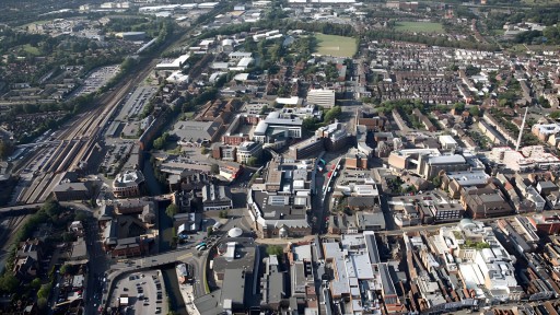 Aerial view of Guildford town center with a mix of commercial buildings and residential areas, highlighting the juxtaposition of historic architecture and modern development, near the railway station and lush green fields.