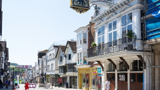 Sunny day on Guildford High Street showcasing historic buildings with traditional facades, including the iconic Guildhall clock, with pedestrians and retail shops lining the road.