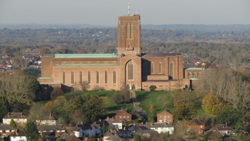 The Cathedral of the Holy Spirit, Guildford, dominating the skyline with its red brick architecture, large central tower, and expansive nave, set against a backdrop of residential houses and autumnal trees.