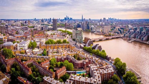 Aerial view of Kensington, London, showcasing its iconic red brick Victorian and Edwardian townhouses, tree-lined streets, and proximity to the River Thames. The image highlights the blend of historic and modern architecture, with the London skyline visible in the distance, including notable landmarks like The Shard. This scenic overview captures the charm and affluence of the Kensington neighbourhood.
