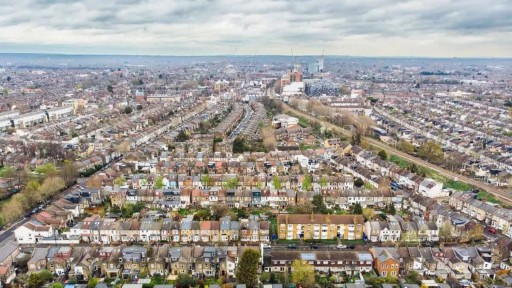 Aerial view of North London's distinctive residential area, showcasing a dense array of colorful terraced houses with varying architectural designs. The foreground features rows of cream and pastel-colored homes with pitched roofs, while the background extends to a horizon of urban development, including construction cranes, underlining the ongoing growth. The image highlights the unique urban tapestry and vibrant community, key points of interest for architectural and urban planning professionals.
