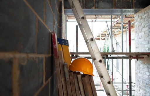 Inside of a construction site, lays pieces of wood against a freshly bricked wall, a yellow hardhat left on the top of the wooden slats, a tall aluminium ladder and peeking through the back is the start of some scaffolding