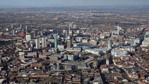 High-angle aerial photograph of the West Midlands urban skyline, showing a mix of modern high-rise buildings and historical architecture, roads, and several green spaces, illustrating the region's urban development and architectural evolution.