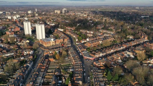 Aerial view of a residential area in West Midlands, showcasing a curved road lined with rows of terraced houses, flanked by taller apartment buildings, and interspersed with greenery, illustrating community planning and architectural diversity.
