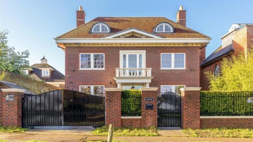 Traditional red brick detached house in Wimbledon with a gated driveway, manicured hedge, and a classic dormer window under a clear blue sky, exemplifying upscale suburban living.