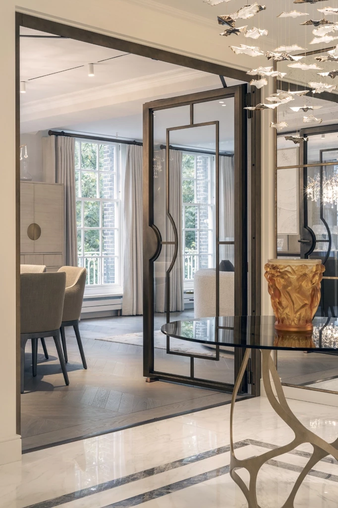 Refined entrance and dining space in a London residential project delivered by Urbanist Architecture, featuring bespoke metal-framed glass doors, a sculptural console with an amber glass piece, elegant marble flooring, and soft natural light filtering through tall sash windows into a calm, contemporary interior.