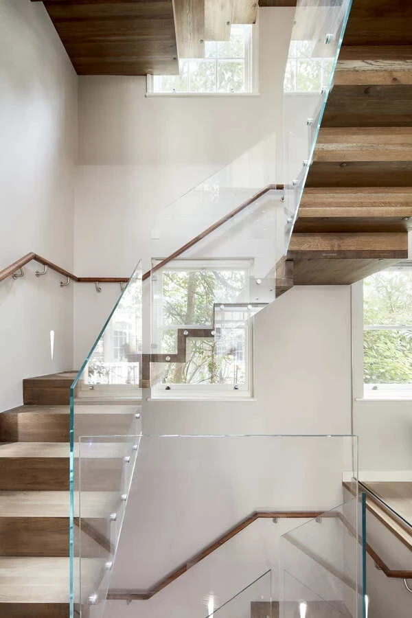 View of a three-storey wooden floating staircase with natural lighting bouncing off of the white walls and travelling through clear glass guard rail 