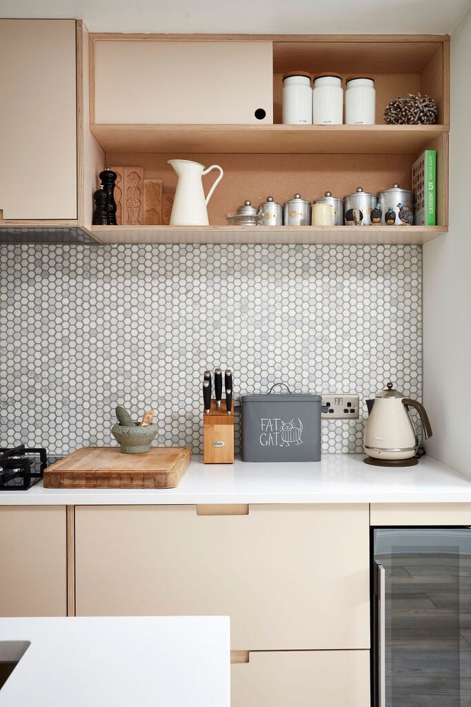 Section of a kitchen work top area with small hexagon grey and white splashback, white countertop, pinky beige cabinets, wine cooler on the bottom right as well as kitchen accessories such as a chopping board, kettle, spices scattered throughout the countertop and shelves