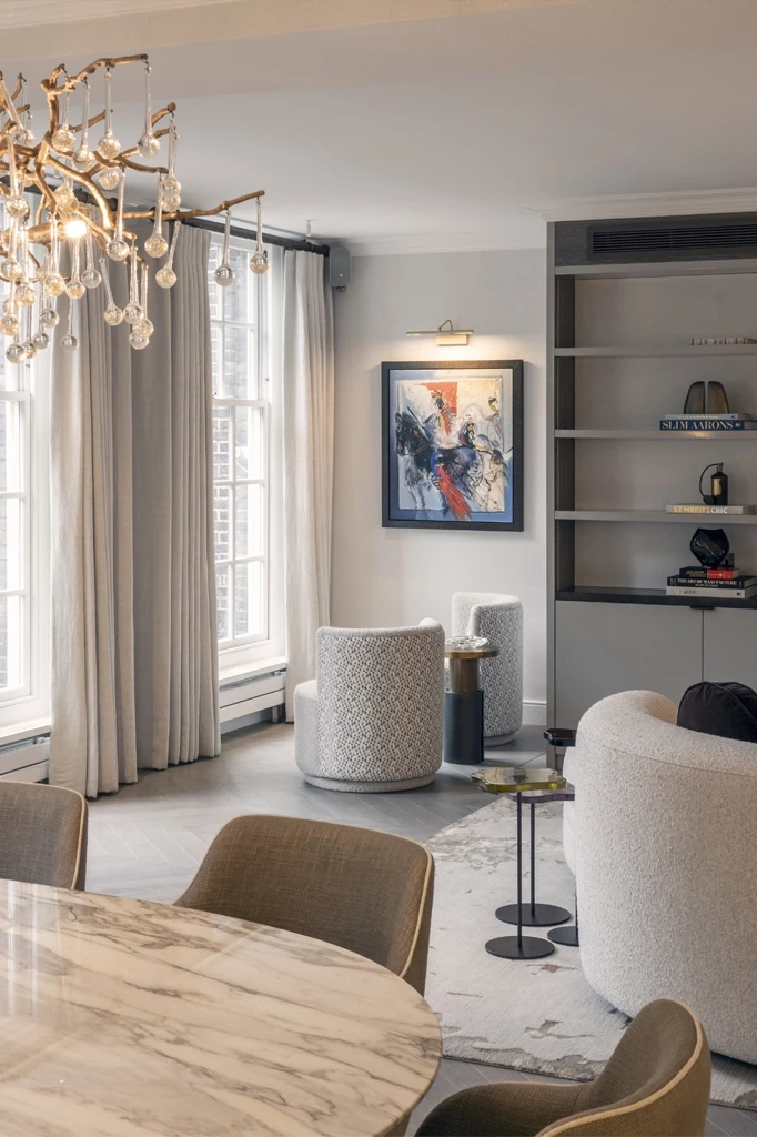 Elegant living area in a London residential project delivered by Urbanist Architecture, featuring large sash windows with soft curtains, a sculptural brass chandelier, a marble dining table, and curved upholstered lounge chairs set within a calm, neutral interior with bespoke shelving and contemporary artwork.