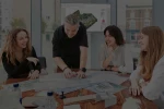 Smiling team of RIBA chartered architects, architectural designers, interior designers and planning consultants collaborating and discussing masterplan and technical drawings printed on paper and on the tv screen behind them