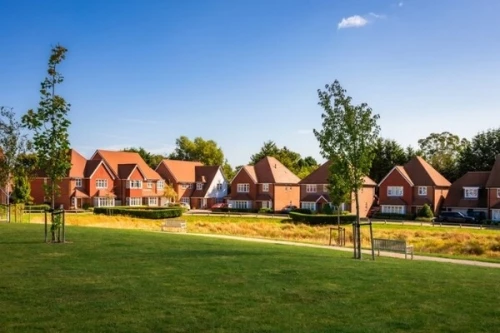A modern housing development featuring red brick homes with terracotta tile roofs situated in Green Belt land. The development includes a mix of two-story houses with white trim and dormer windows, set back from a large grassy area with young planted trees.
