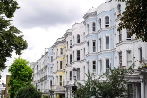 Pastel-painted Victorian terraces in London framed by summer trees, handsome but built for cooler climates, now needing heat-resilience retrofits.
