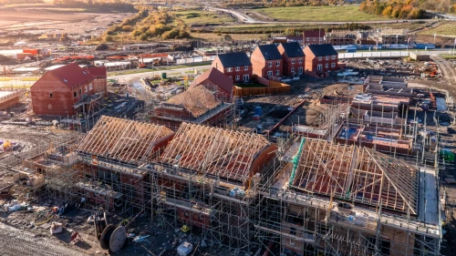 Aerial view of a large housing development under construction, showing rows of new brick houses with timber roof frames and scaffolding, surrounded by open countryside and farmland.