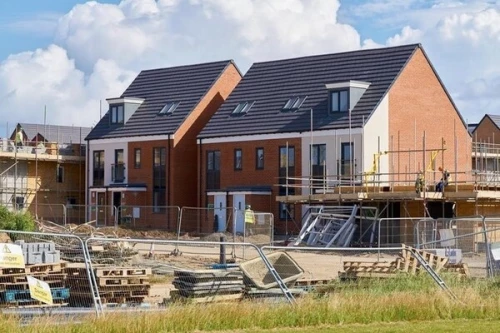A UK housing development under construction showing new-build red brick homes at various stages of completion. Finished three-storey houses stand alongside partially built structures with scaffolding, while the foreground shows construction site fencing, stacked pallets, and overgrown grass, illustrating the challenges housebuilders face in delivering quality homes amid viability pressures that threaten the government's target of 1.5 million new dwellings.