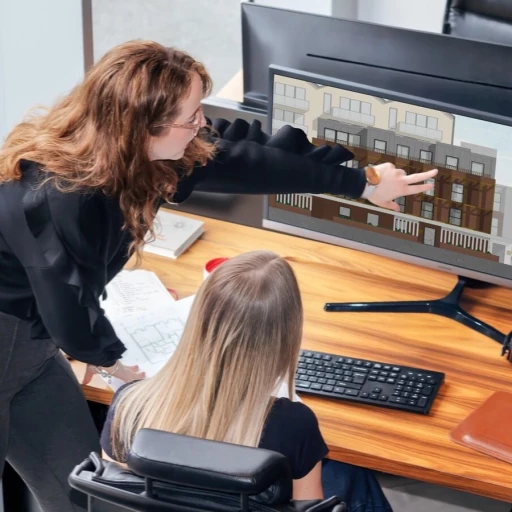 Two architects collaborating at a wooden desk in a modern office setting at Urbanist Architecture, a small RIBA chartered architecture firm in London. One woman with curly brown hair and glasses stands while pointing at architectural elevation drawings displayed on a large monitor, while another woman with straight blonde hair sits at the desk. The screen shows detailed building facade designs with multiple stories and varied window configurations. Papers and documents are spread across the wooden desk surface, along with a black keyboard, demonstrating the collaborative design process typical of professional architectural practice.