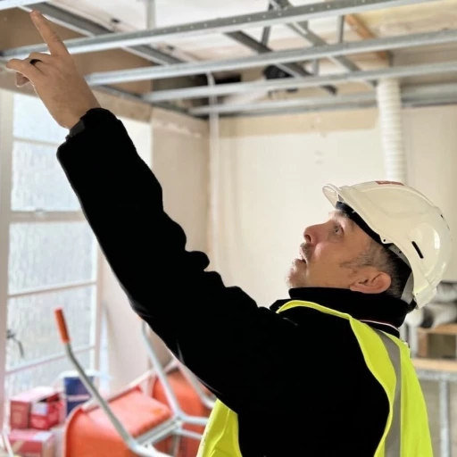 Chartered architect in hard hat and high-visibility jacket inspecting suspended ceiling framework on site, demonstrating agile project management in construction practices. This hands-on site supervision exemplifies the director-led, hands-on involvement from first sketch to final handover approach used by chartered architects in London for comprehensive project management. The image showcases active project management for construction, where the chartered architect maintains direct oversight of building works to ensure quality control and adherence to architectural specifications.