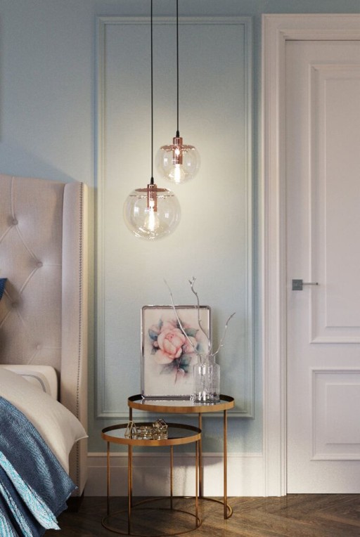 Photograph of brass circular night tables next to a luxurious padded cream bed with high headboard bed frame, copper and glass pendant lights and a baby blue wall and bright white door