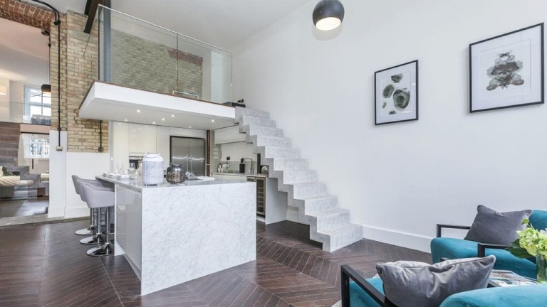 Modern renovated apartment in a Grade II-listed building featuring a sleek marble kitchen island with bar seating, a minimalist staircase leading to a glass-enclosed mezzanine, and a comfortable seating area. The design integrates exposed brickwork and contemporary finishes, showcasing a blend of historical charm and modern elegance.