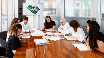 Team of architectural designers, interior designers, graphic designer and planning consultants around a shiny wooden table discussing Green Belt project with various designs printed and spread on the table as well as on the tv screen behind them